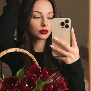 А girl taking a mirror selfie indoors, medium close-up shot with a slight high angle and three-quarter framing; right hand raised and bent at the elbow, holding a metallic gold smartphone with a triple-lens module near cheek level, fingers wrapped around 