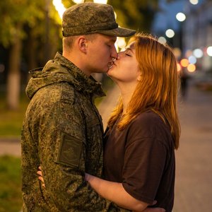 На фотографии должна быть изображена трогательная сцена нежной любви и эмоциональной близости. Молодой человек в военной форме, вероятно, в армейской форме, обнимает и целует девушку. Он стоит чуть слева, что добавляет фотографическому и атмосферному эффе