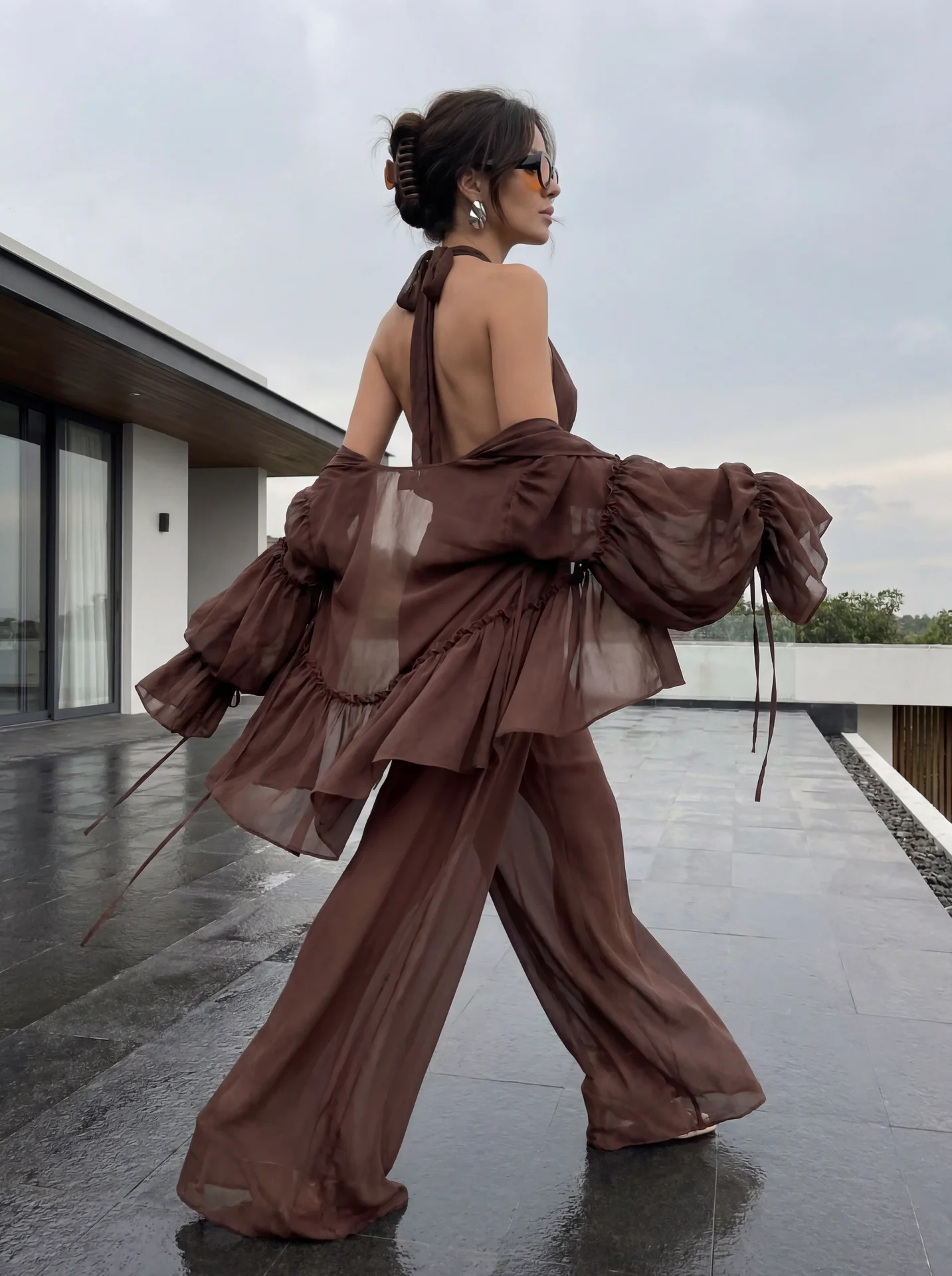 Ультрареалистичное luxury feminine fashion фото точно как на референсе: девушка снята со спины и немного сбоку в движении, она идет вперед по темной современной террасе. Одна нога делает широкий шаг вперед, другая остается сзади, бедра мягко раскачиваются