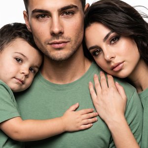 An ultra-realistic, close-up studio portrait, intimate and emotional, with no alterations to facial features. Three people fill the frame from edge to edge. The man is centered, looking forward, calm and confident. The woman has her head on his shoulder, 