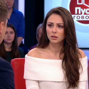 Young woman sitting in the guest chair on a dramatic Russian television talk show similar to “Пусть говорят,” filmed by a live studio broadcast camera. She is seated under bright studio lights, looking emotional and slightly surprised, with a serious expr