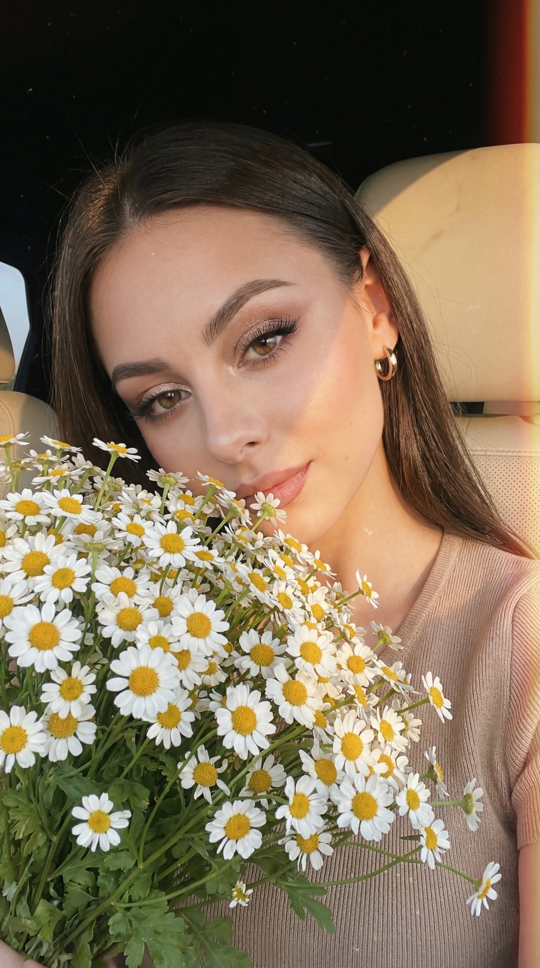 A girl takes a selfie in close-up, slightly tilted forward-on the right at eye level (85 mm, f/1.8), in the car at the window, lighting is a sunset fan, sunset, rays fall beautifully on objects; a bouquet of white daisies is held up to her face, flowers d