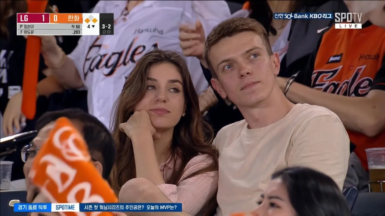 Create subtle natural movement for the main character positioned in the center of the frame, as if captured live by a real stadium broadcast camera during a crowded baseball game. The character should naturally react to the match with realistic eye moveme