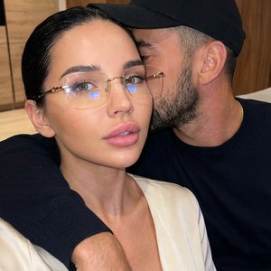 Интимный портрет пары крупным планом: женщина на переднем плане и мужчина позади нее. У женщины  волосы зачесаны назад, она носит очки без оправы с золотыми вставками и светлый верх, возможно, белую или кремовую блузку с намеком на декольте. У нее пухлые 