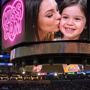 Young woman sitting at a Lakers playoff game shown on the giant arena screen during a kiss cam moment. Camera is closer to the jumbotron so her face is clearly visible and fills most of the frame. She is leaning in and affectionately kissing a cute young 