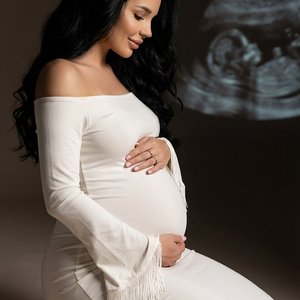 Фотореалистичная студийная maternity\-фотография в мягком кинематографичном editorial\-стиле\. Беременная женщина с длинными блестящими густыми волнистыми волосами сидит на коленях \(на пятках\) на светлом бесшовном фоне, профиль влево\. Вертикальный кадр