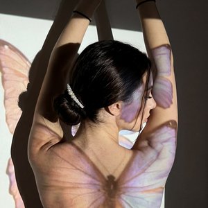 A moody, vertical 9:16 photo taken on an iPhone in a dimly lit room at night — likely a bedroom or studio space with plain walls and minimal decor. The subject is a young woman seen from behind, standing close to a wall where projected light casts dramati