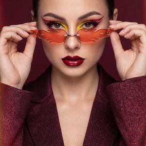 A girl, medium close-up shot, eye-level camera angle, professional high-fashion studio photography. Slender figure. Shoulders squared forward, head tilted slightly down. Gaze locked piercingly directly into the camera over the rims of lowered sunglasses. 