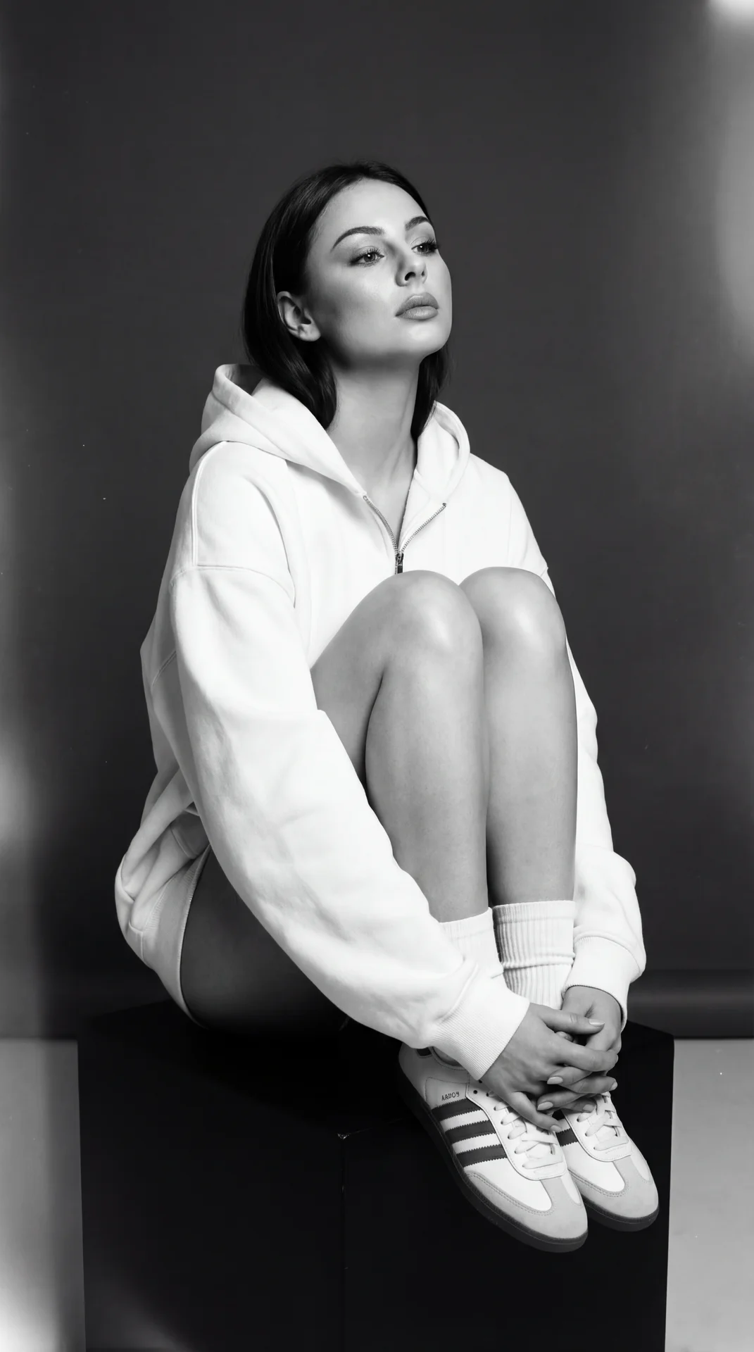 A girl — vertical professional portrait, full seated crop from top of head to shoes, camera positioned at slight low‑to‑eye level (mild upward tilt), shallow depth of field for subject separation.

Pose and hands:
⦁ Sitting on a low black cube, torso lean
