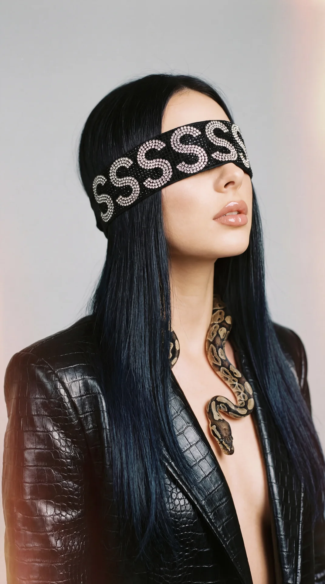 A girl is posed in a three-quarter profile facing to the right, with her head tilted slightly upward. Her eyes are completely covered by a wide, black blindfold adorned with sparkling rhinestones arranged to form a repeating “S” pattern, adding a striking
