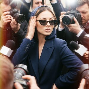 A girl in a tight dress in close-up, body slightly tilted forward, standing right in the middle of a dense crowd of journalists; chin slightly raised; head slightly to the side; gaze glances away; mouth closed with relaxed neutral lips; one hand is raised