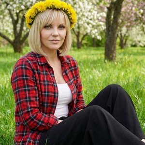 Девушка сидит в зеленой траве , сзади деревья цветущие. На голове у нее венок из одуванчиков. Одета в клетчатую рубашку, под рубашкой белый топ .и черные штаны"полацио". Легкий макияж