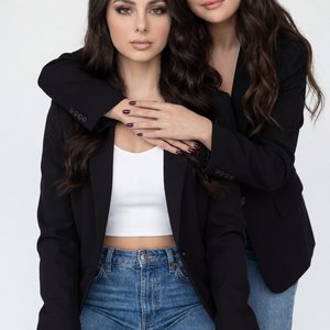 Two women in a minimal white studio, professional portrait, medium shot, eye-level straight-on; front subject seated on a white cube, knees slightly apart, hands relaxed on thighs, serene closed-lip expression, direct gaze; rear subject close behind, lean