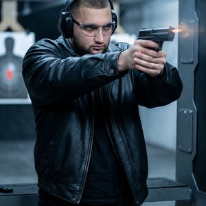 Создай изображение мужчины, стреляющего из пистолета в помещении для тренировочной стрельбы. Он виден по пояс, стоит в устойчивой стойке, обеими руками держит пистолет, направленный в сторону мишени. На нём чёрная футболка черные джинсы и чёрная кожаная к