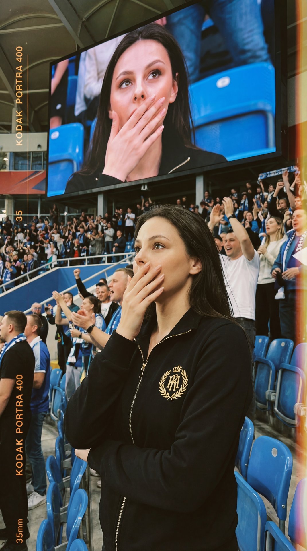 A girl stands in a crowded indoor stadium, captured in a professional medium shot with a slightly elevated camera angle that includes both her and the large screen behind her displaying her close-up. She is positioned slightly off-center among seated and 