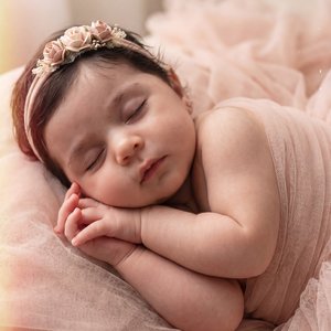Newborn baby sleeping in an indoor studio, swaddled in sheer blush‑pink tulle; floral headband with small blush roses; lying on side with torso gently curled, head slightly tilted toward camera and a touch to camera right; forearms bent and stacked under 