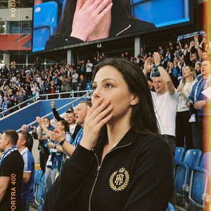 A girl stands in a crowded indoor stadium, captured in a professional medium shot with a slightly elevated camera angle that includes both her and the large screen behind her displaying her close-up. She is positioned slightly off-center among seated and 