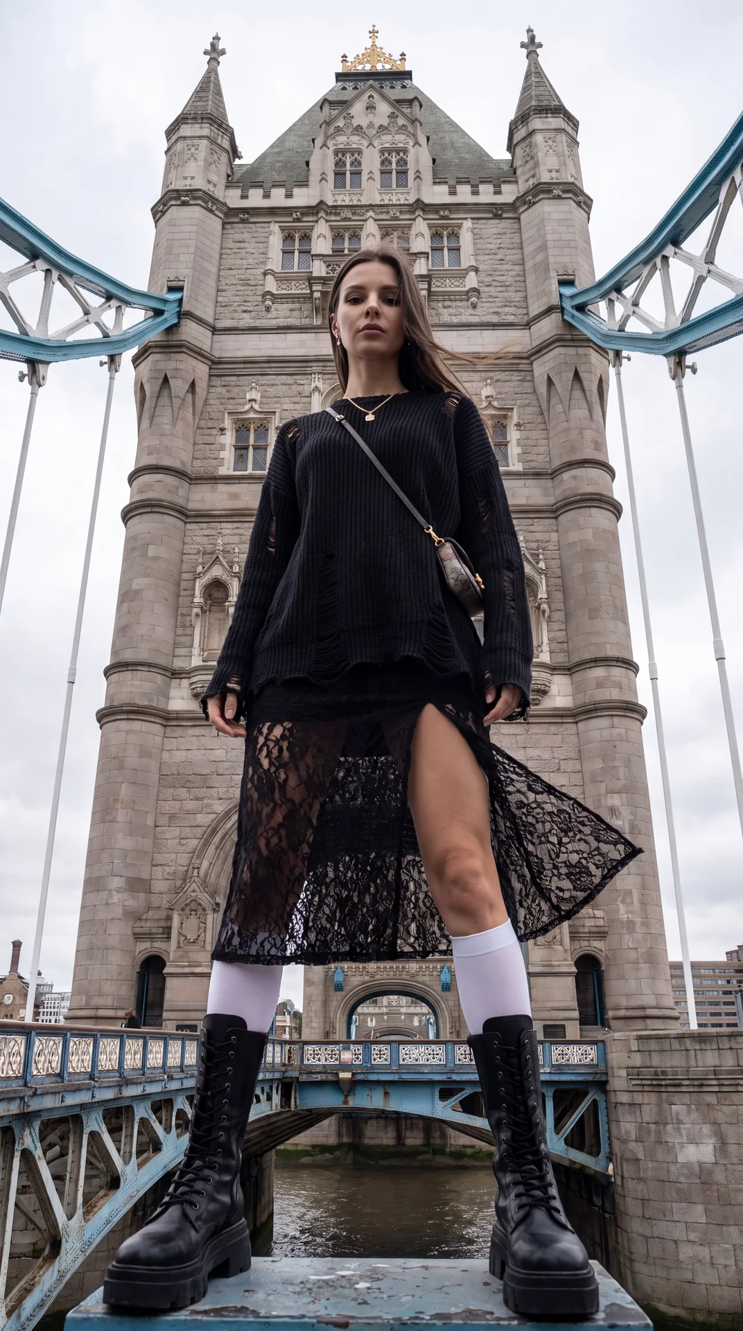 A tall model is standing by one of the towers of Tower Bridge. Outfit:
an oversized black top with a distressed look,
a black lace skirt with a slit, white knee-high socks, and black
platform boots. Accessories: a small crossbody bag, a chain necklace wit