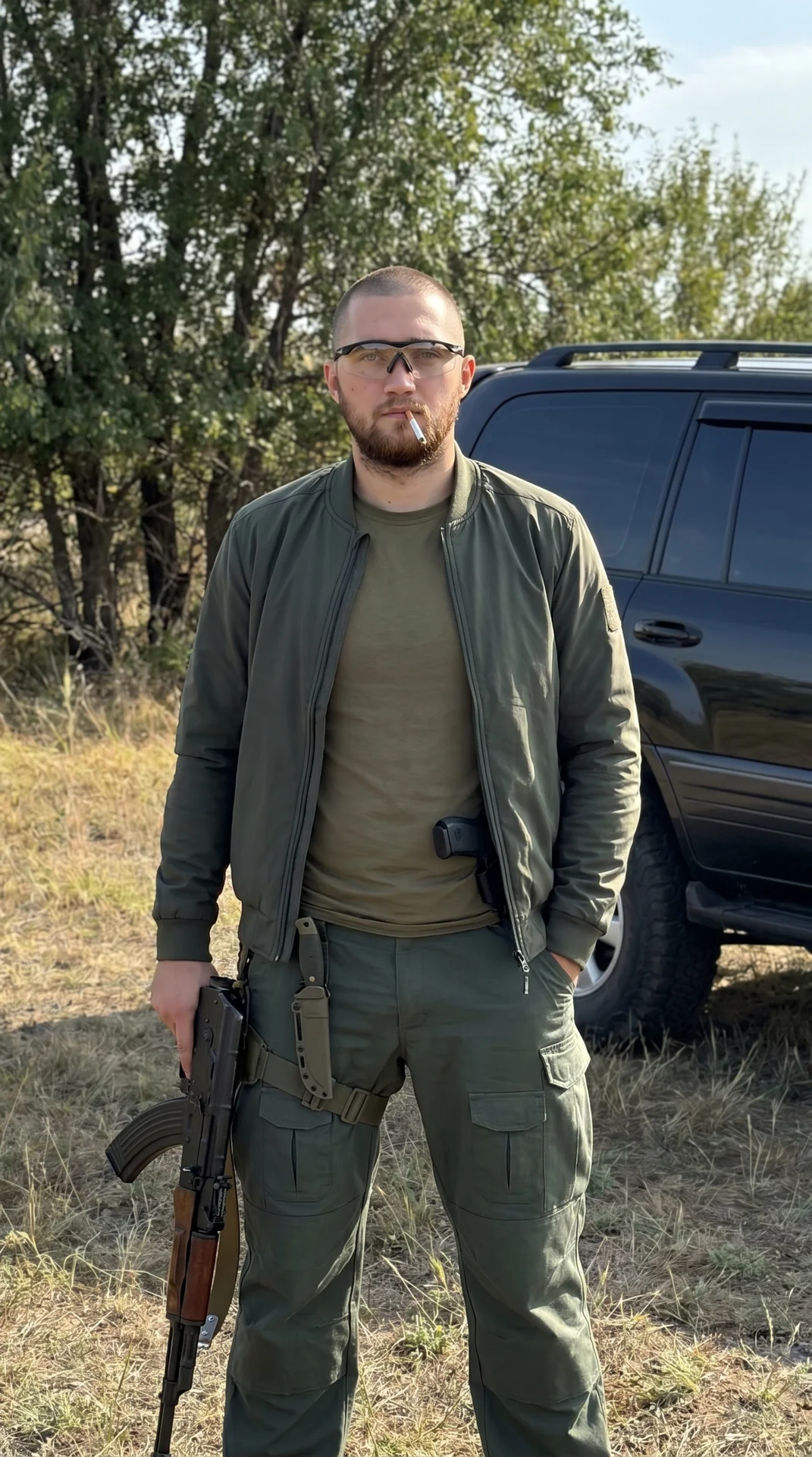 Высококачественное фото молодого мужчины в стиле military. На нем приталенная темно-зеленая (болотного цвета) куртка-бомбер без воротника, надетая поверх оливковой базовой футболки, и широкие темно-зеленые тактические штаны с карманами, на правом бедре та