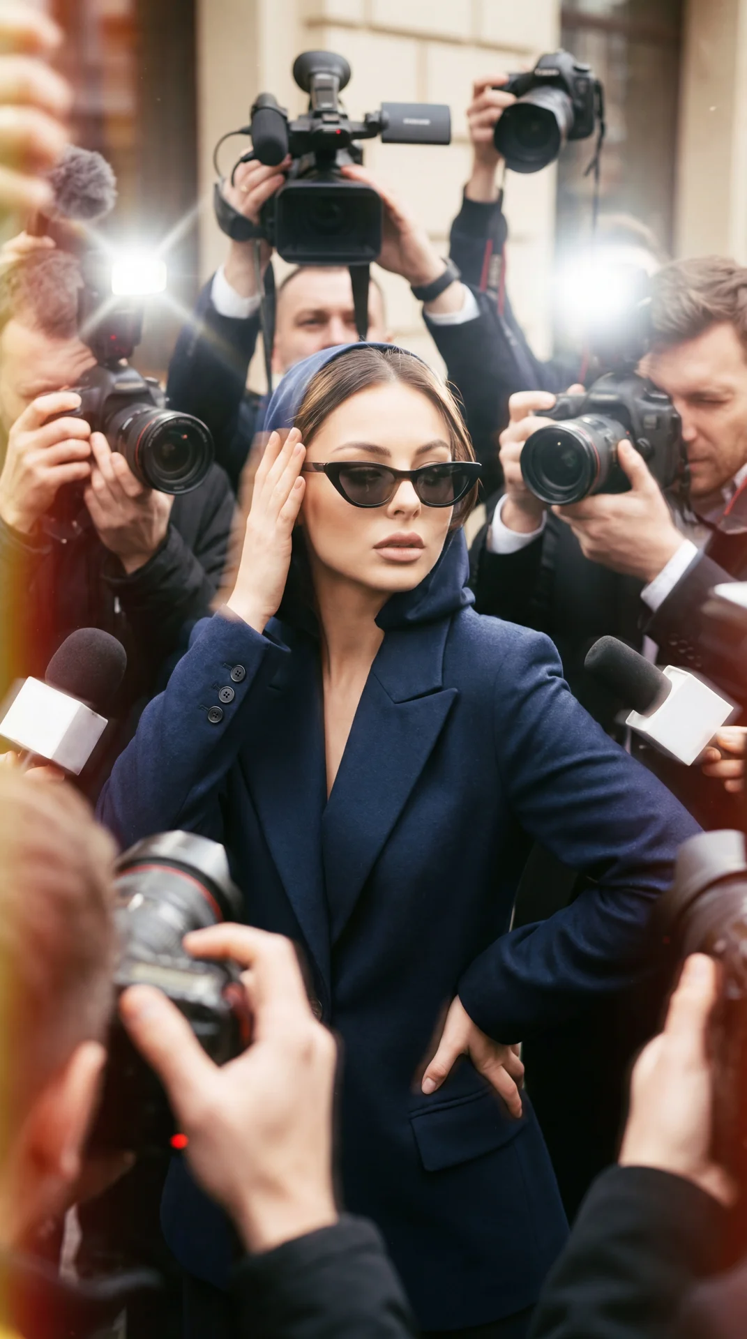 A girl in a tight dress in close-up, body slightly tilted forward, standing right in the middle of a dense crowd of journalists; chin slightly raised; head slightly to the side; gaze glances away; mouth closed with relaxed neutral lips; one hand is raised