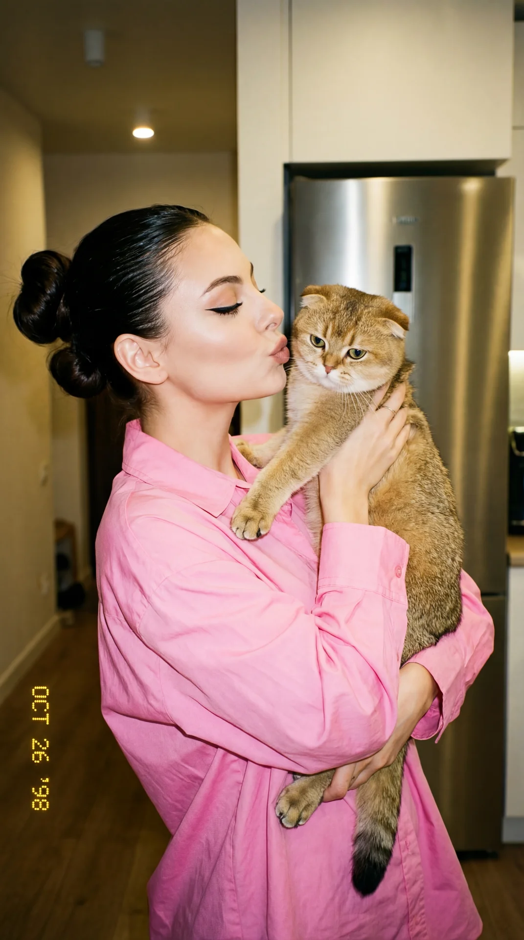 A girl with slicked‑back hair gathered in two voluminous buns, dressed in a spacious bright pink cotton poplin button-down shirt, stands in a room near a stainless steel refrigerator; the body is three-quarters turned to the camera to the right, a straigh