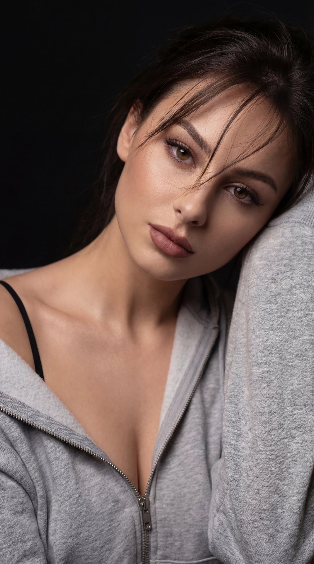 A girl is posed in a close-up portrait against a deep black background, creating an intimate and moody atmosphere with dramatic lighting that emphasizes highlights and shadows on her skin. She is leaning slightly forward, with her head tilted gently to th