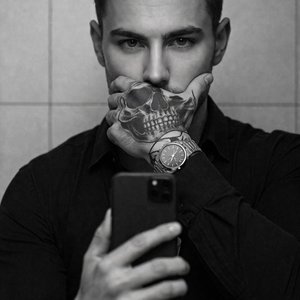 A men in a monochrome, high-contrast mirror selfie, tight medium close-up at eye level, professional-camera look (50mm equivalent, f/1.8, shallow depth, crisp focus, soft directional light), indoors against matte ceramic tiles. Pose: torso facing camera w