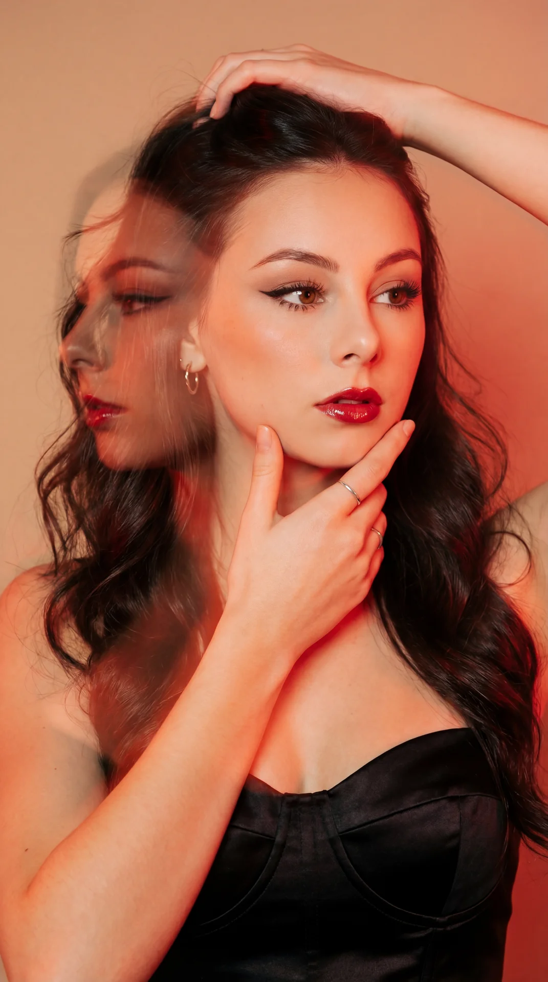 А girl in a studio, vertical tight medium close-up at eye level, shot on a professional camera with an 85mm lens and shallow depth of field; double exposure overlays a semi-transparent offset profile to the left; warm red/orange gel wash, light-leak glow,