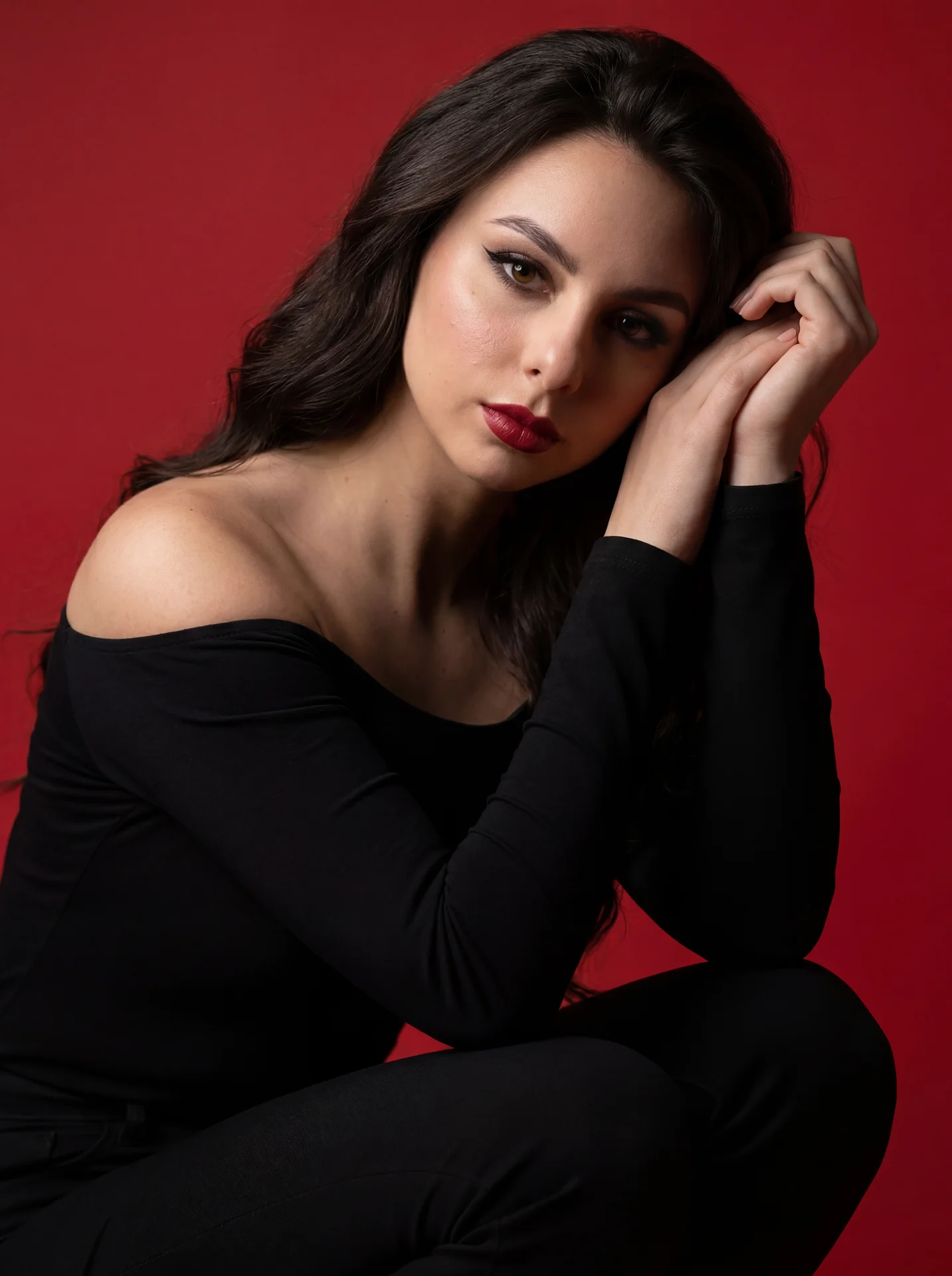 Professional studio portrait, tight three-quarter shot at eye level on a full-frame 85mm, woman in a matte black off-shoulder long-sleeve top and black pants, crouched with one knee raised and torso slightly forward; right forearm vertical resting on the 