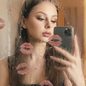 A girl is standing behind a fogged glass surface covered with water droplets and streaks, which partially obscure her figure and face, creating a soft, blurred effect. She holds a smartphone in her right hand raised to a mid-chest level, capturing a mirro