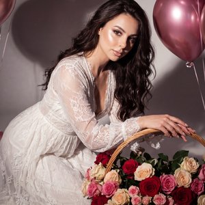 Создай не меняя черты лица 
Соотношение сторон 9:16
молодая женщина в белом лёгком платье, присевшая на колени и слегка наклонившаяся вперёд, обе руки положены на большую плетёную корзину с пышным букетом роз (красные, розовые, кремовые), рядом металличес