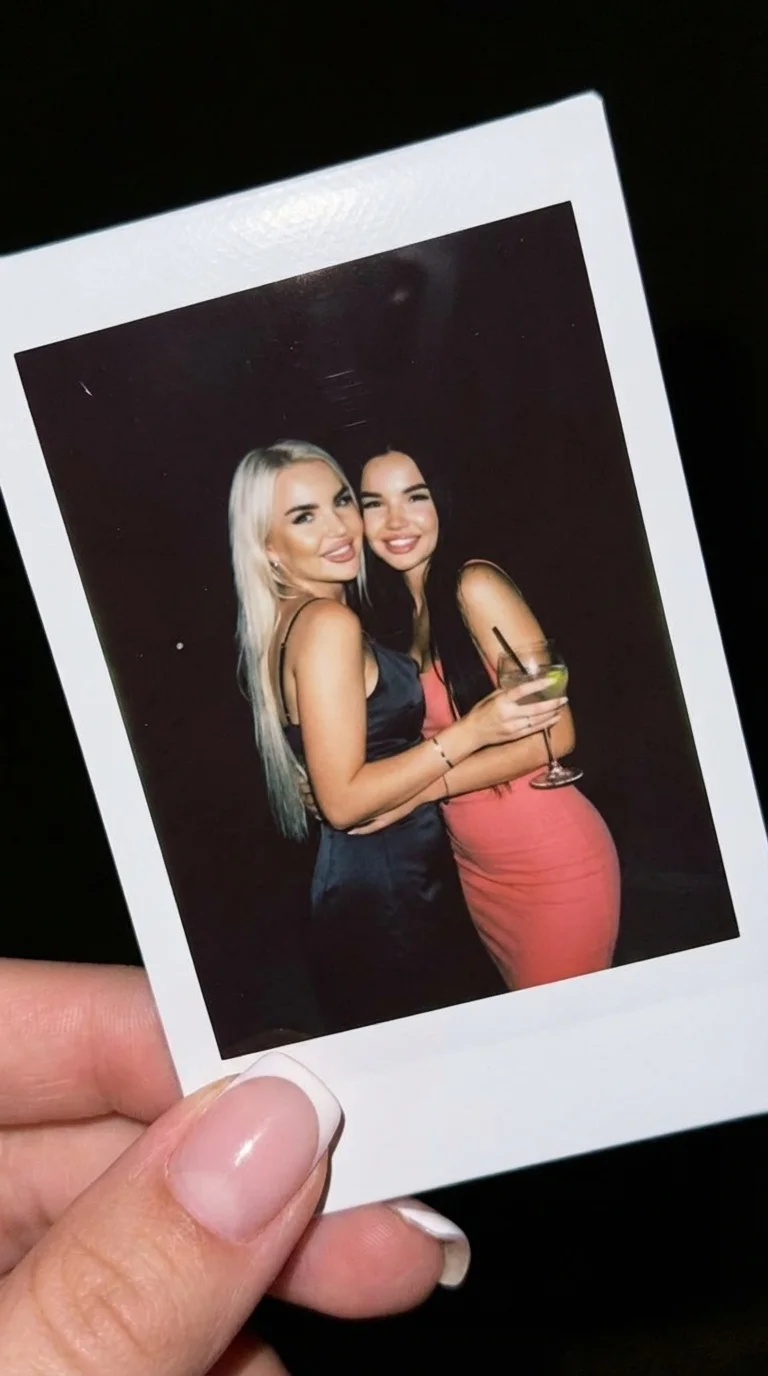 5. A photograph of two women, each held in a hand-held snapshot against a dark background, is slightly tilted, the composition is diagonal, and the main focus is on the Polaroid frame itself. In the image, the women stand close together, shoulder to shoul