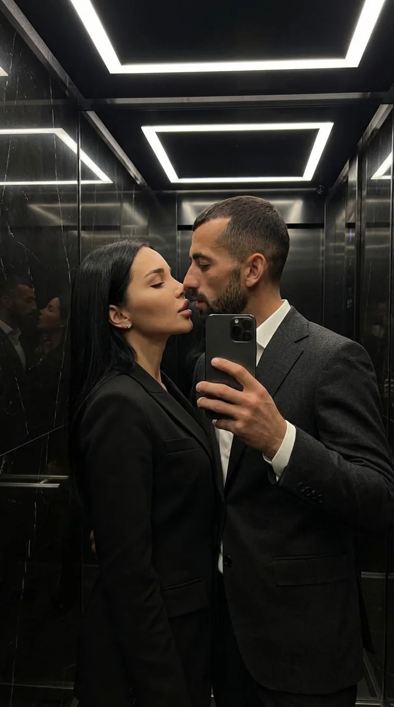 A luxurious, intimate mirror selfie inside a dark elevator with reflective black walls and soft linear ceiling lights. A well-dressed couple stands extremely close, almost touching lips.
The man wears a tailored dark wool suit with a crisp white shirt, ho