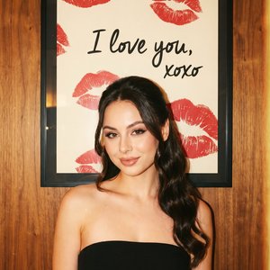 The image features a person standing in front of a framed artwork on a wooden wall. The individual is wearing a strapless black dress and has long, wavy dark hair cascading over one shoulder. The expression on their face is pleasant and relaxed.

The artw
