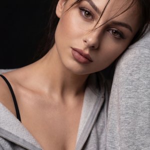 A girl is posed in a close-up portrait against a deep black background, creating an intimate and moody atmosphere with dramatic lighting that emphasizes highlights and shadows on her skin. She is leaning slightly forward, with her head tilted gently to th