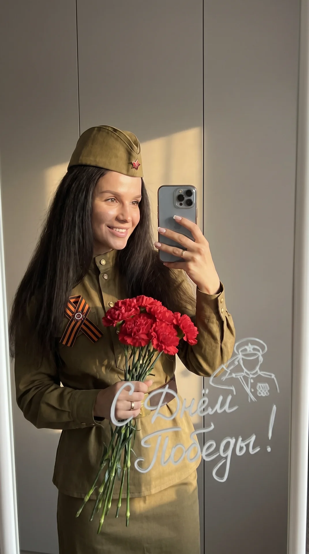 Девушка делает фото в зеркало на телефон. Она одета в советскую военную форму и на голове советская военная пилотка, на груди приколота Георгиевская ленточка. Одной рукой она держит букет красных гвоздик Фон нейтральный, стены серые, создавая спокойную ат