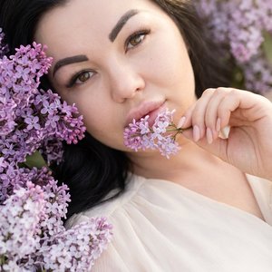 Не изменяй лицо, черты лица.
Крупный план женщины в окружении сиреневых и светло-лиловых соцветий сирени, стиль романтической, мягкой фотографии. Поза: женщина лежит или полулежит среди цветов, держит веточку сирени возле рта; плечи и часть груди в нежной