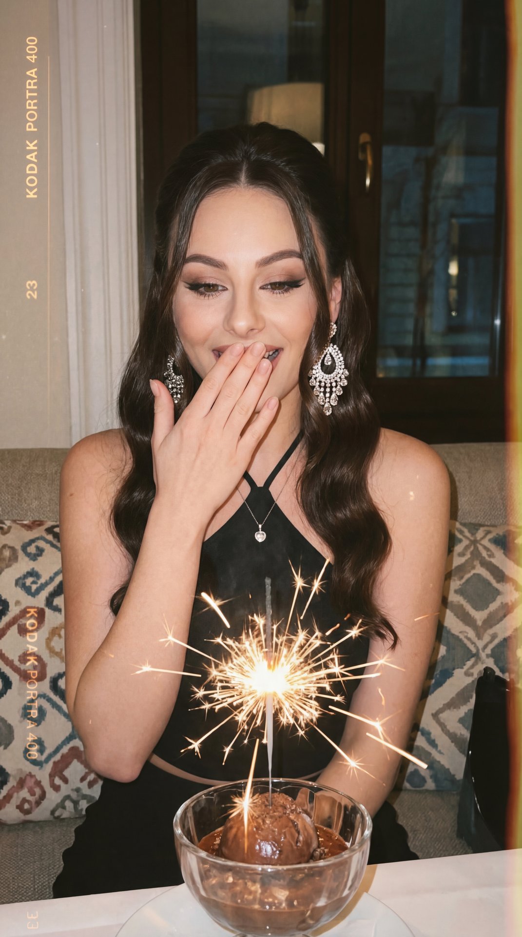 The image captures a joyful moment where a woman is reacting with delight. She is dressed elegantly in a black outfit and accessorized with sparkling, ornate earrings. Her long dark hair complements her look, and she wears a subtle necklace with a pendant