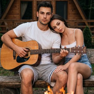 Это снимок, похожий на кадр из фотосессии, сделанный на природе возле деревянного дома в форме буквы "А". Внизу кадра виден горящий мангал с дровами, пламя от которого немного освещает передний план. Атмосфера теплая и непринужденная.

На переднем плане с