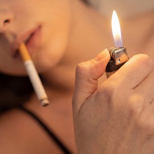 Модель в чёрной майке с тонкими бретелями, на бежевом диване, мягкий тёплый рассеянный свет — поменять позу: сидит полуобернувшись, правая рука поднята ближе к объективу; макро-съёмка 1:1 с очень малой глубиной резкости, фокус на пламе зажигалки, металлич