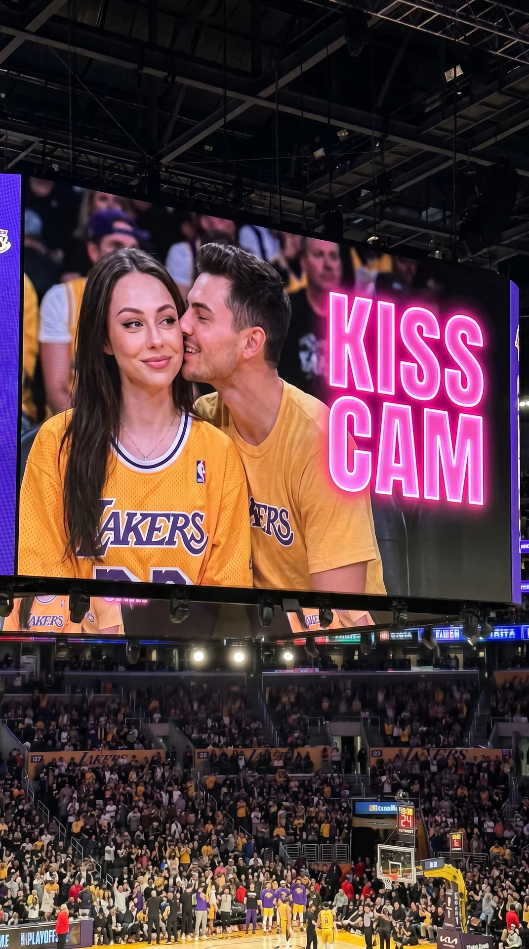 Ultra-realistic NBA jumbotron kiss cam screenshot, identity preserved exactly from reference image.

Young woman sitting at a Lakers playoff game shown on the giant arena screen during a kiss cam moment. Camera is closer to the jumbotron so her face is cl
