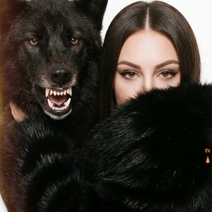 A girl is positioned closely alongside a large black wolf with its mouth open in a fierce snarl, revealing sharp teeth and textured gums. Her face is partially obscured by the thick, dark fur of what appears to be a luxurious black fur coat or stole that 