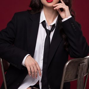 Professional studio fashion portrait: a woman sitting on a vintage metal chair against a dark red seamless background; medium shot, slightly lowered viewing angle, three‑quarter pose; torso tilted forward, one knee raised to the lens; left forearm resting