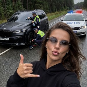Не меняя внешности женщины с фото создай селфи кадр на фронтальную камеру iPhone, 
сцена происходит в пасмурную погоду на мокрой асфальтированной трассе сразу после дождя, небо отличается серостью, влажный воздух,по обе стороны густой хвойный лес и линная