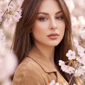 Professional close-up portrait of a woman among blooming cherry blossoms, shot on a full-frame camera with an 85mm lens at f/1.8, ISO 100, 1/500s; eye-level, straight-on angle, portrait orientation, center-framed; pose: shoulders relaxed and facing the ca