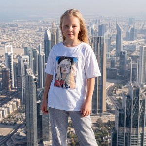 Фотореалистичное изображение: девочка 7 лет из фото, внешность и черты лица максимально соответствуют исходному фото-референсу, стоит на крыше многоэтажного здания днём при солнечной погоде. На ней широкие серые спортивные штаны, белая оверсайз майка с пр
