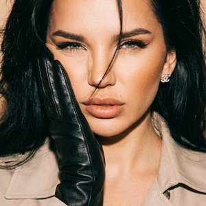 transfer the face and human parameters from my reference photo, preserve identity 100%. Ultra-realistic close-up, centered, eye-level. Black leather glove pressed to cheek. Expression intense feline squint, lips softly parted. Hair voluminous, brushed bac