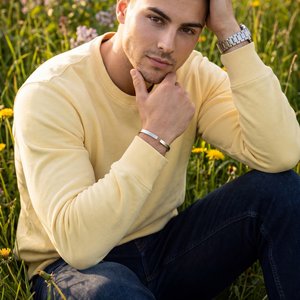 Professional photo, medium shot, slight high-angle (10–15°) looking down; model seated in a lush wildflower meadow (tall green grasses with yellow and lilac blooms); golden-hour natural light from front-left, warm tone, shallow depth of field, soft bokeh,