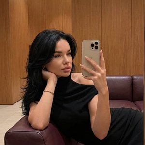 Ультрареалистичное фото 9:16, снято на iPhone, зеркальное селфи. Камера находится на уровне лица, кадр плотный, с лёгким наклоном, создаёт ощущение личного, не постановочного момента.

Сцена — минималистичный интерьер с тёплой атмосферой: стены с деревянн