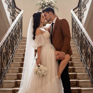 Elegant wedding editorial on a grand double staircase, centered composition with leading lines from ornate wrought-iron railings, vertical portrait, medium-full shot from a slightly low upward angle, full‑frame 35mm lens at f/1.8, soft natural backlight f
